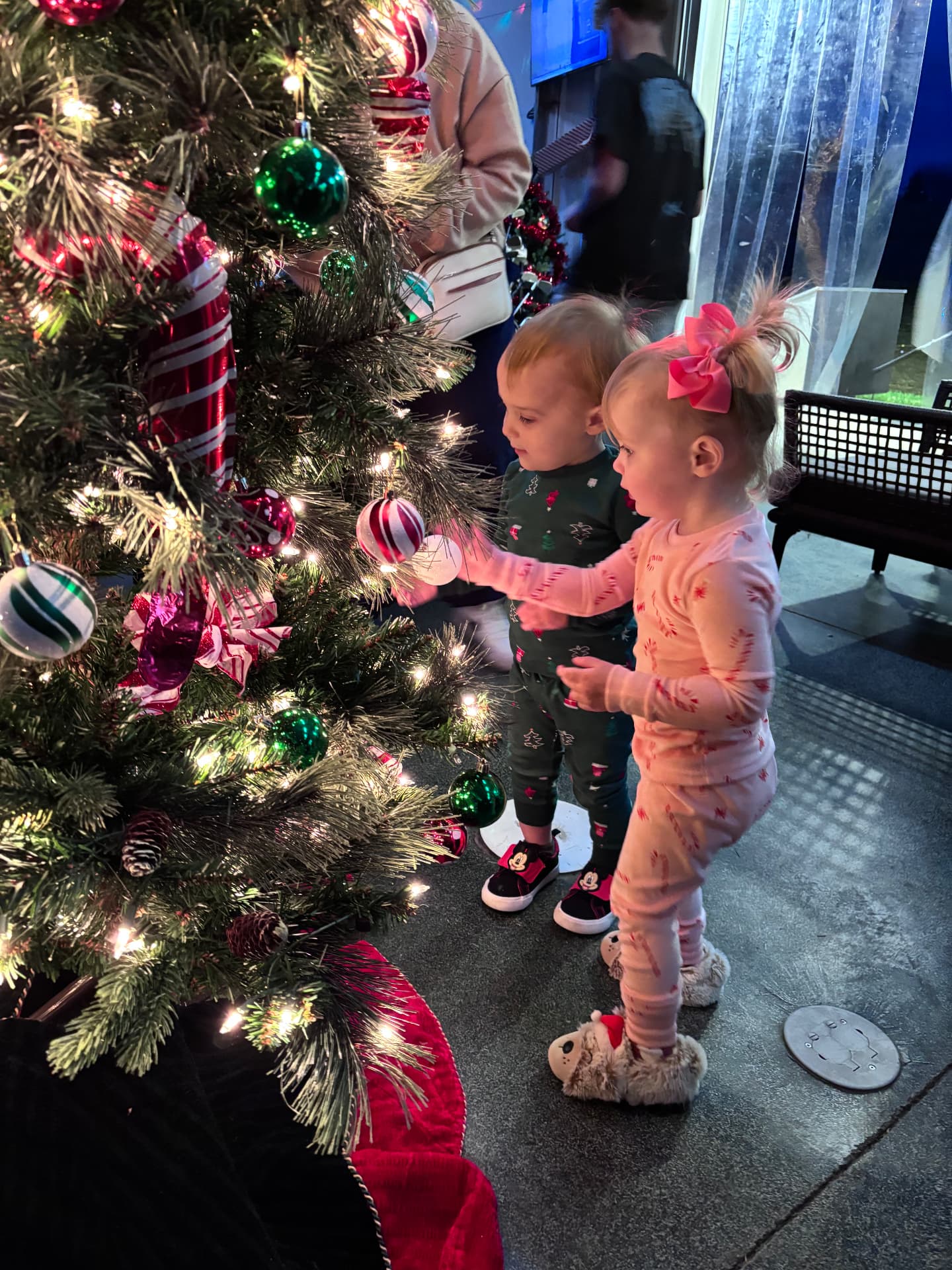 Two toddlers in pajamas touching ornaments on a Christmas tree at The Flight Zone